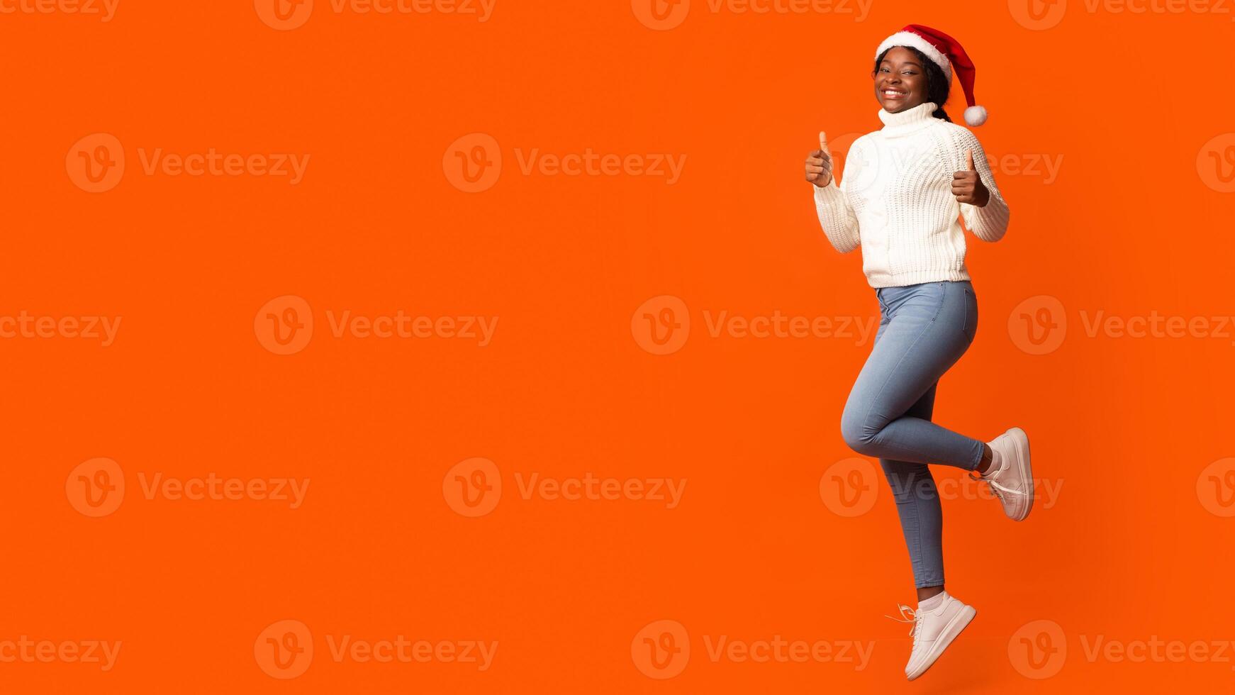 Holiday Offer. Joyful Black Santa Lady In Christmas Hat Jumping And Showing Thumb Up Gesture, Recommending Something, Having Fun On Orange Background In Studio, Cheering Xmas And New Year, Copy Space photo