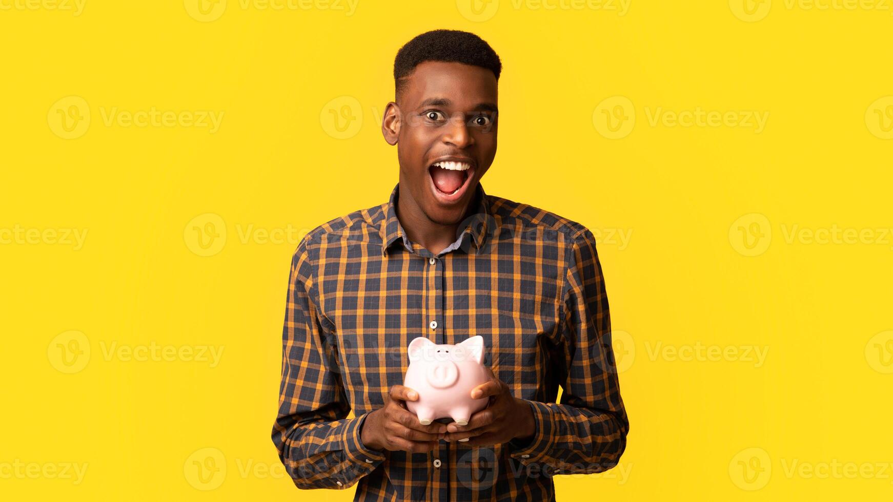 Wise Saving. Portraif Of Funny Excited Black Guy With Piggy Bank In Hands Posing Over Yellow Studio Background, Emotional African American Man Enjoying Economy, Planning His Budget And Investments photo