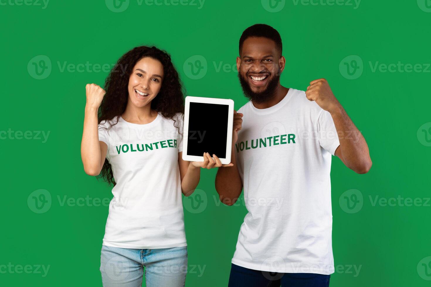Two Multiethnic Volunteers Showing Digital Tablet Empty Screen Gesturing Yes Celebrating Success Standing Over Green Studio Background, Smiling To Camera. Activists Recommending Volunteering Website photo