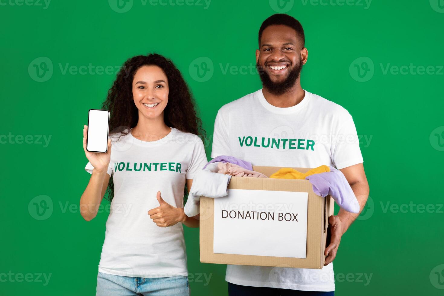 Two Diverse Volunteers Holding Donation Box Showing Smartphone With Blank Screen Picking Clothes For Second-Hand Use Standing Over Green Studio Background, Gesturing Like. Charity And Contribution photo