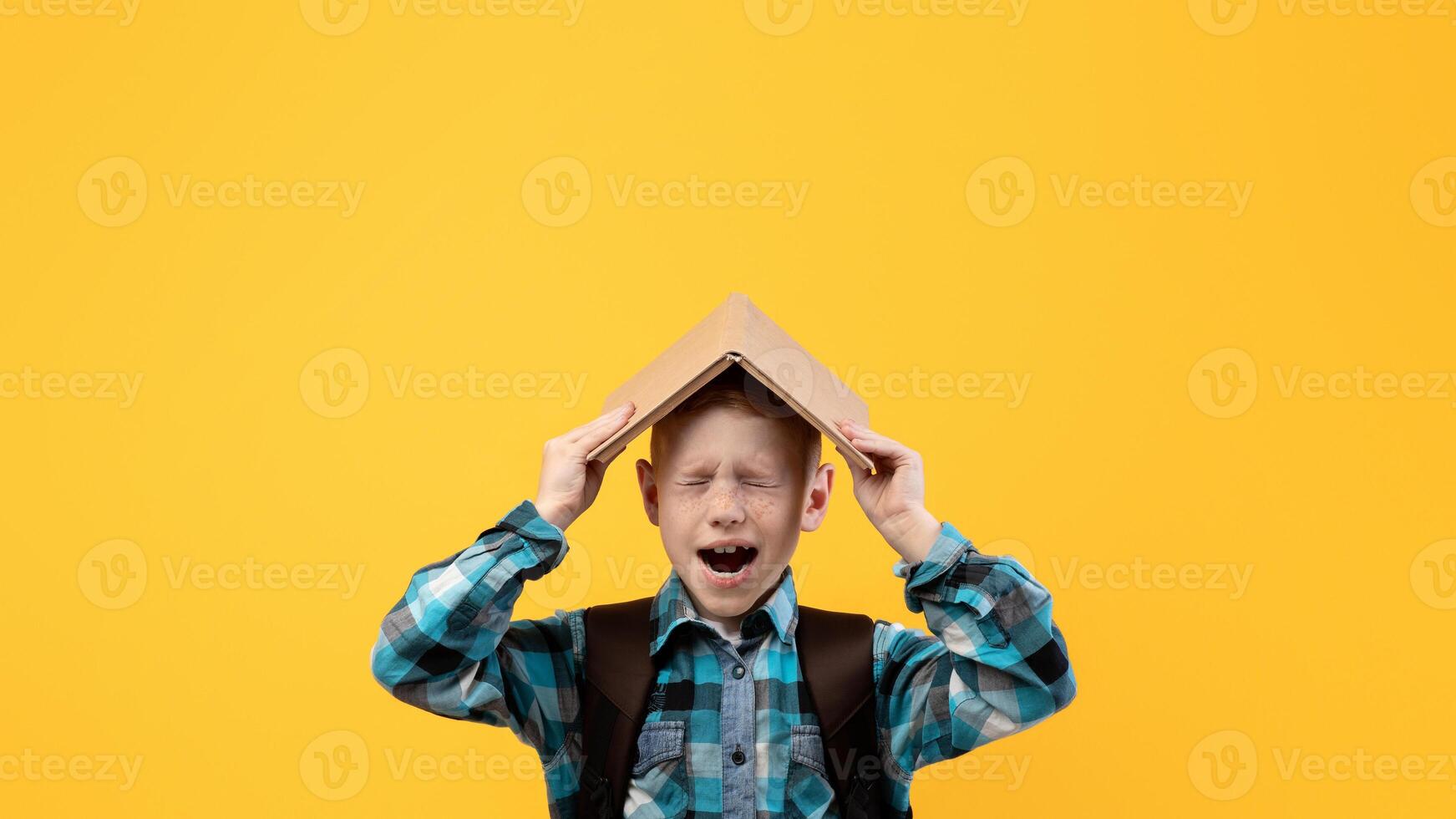 Stressed schoolboy with backpack covering head with book and yelling on yellow studio background, got tired of studying, need for rest, copy space. Ginger boy holding book on his head and screaming photo