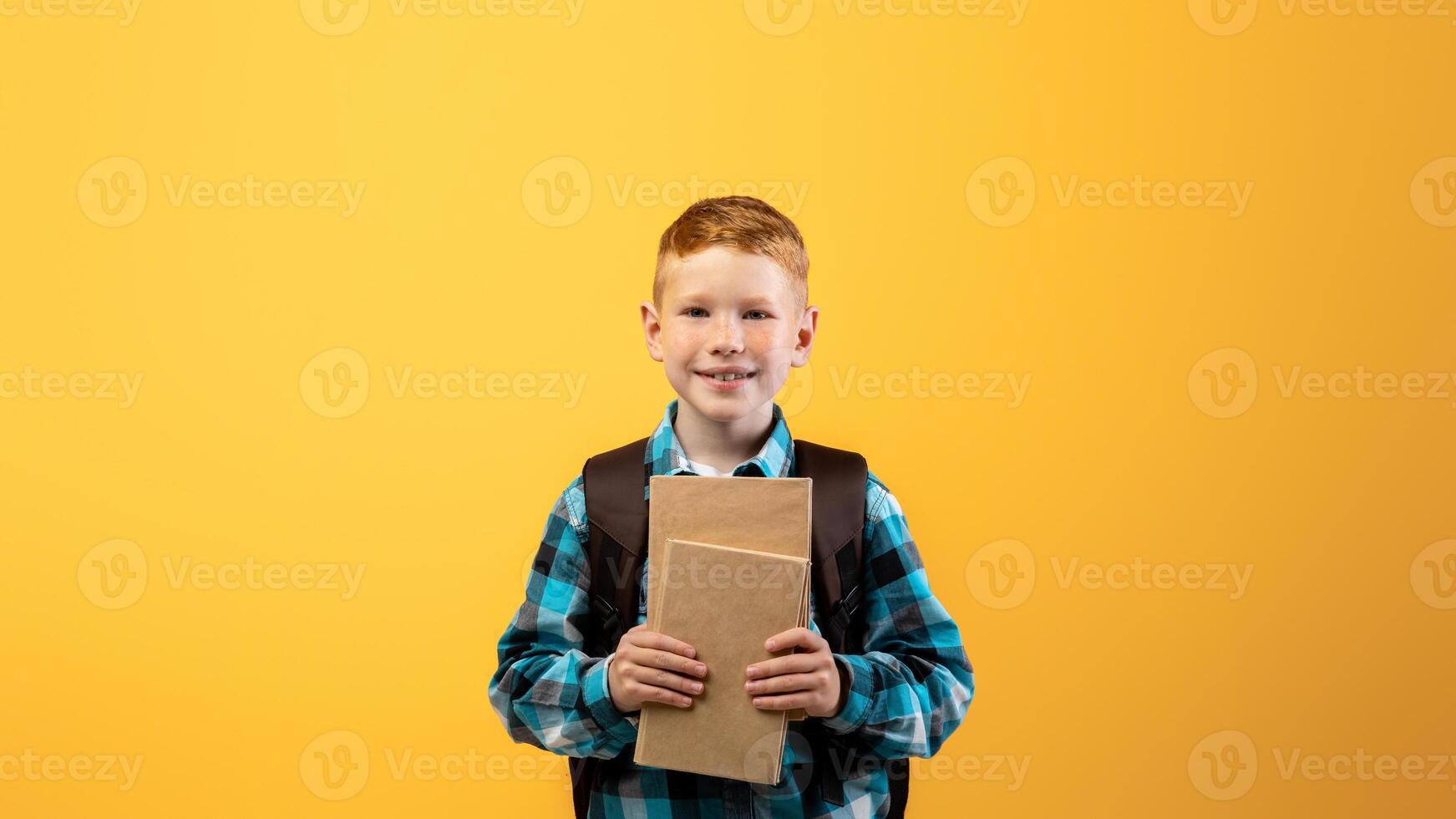 Cheerful boy schooler with backpack holding heap of books, yellow studio background, copy space. Happy boy going to school, holding books, enjoying his school days. Kids education concept photo
