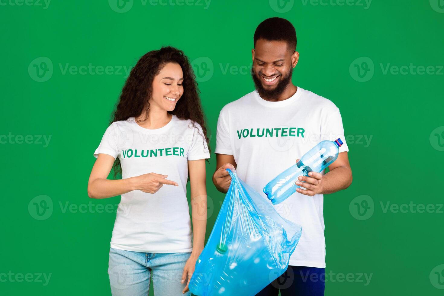 Don't Litter. Male And Female Multiethnic Volunteers Putting Used Plastic Bottle To Blue Garbage Bag Standing Over Green Studio Background. Keep Environment Clean, Ecology And Volunteering Work photo