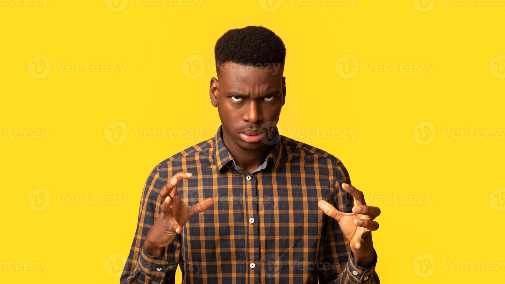Portrait of annoyed black guy in checkered shirt emotionally gesturing, raising hands and rolling eyes with rage, feeling angry and stressed while standing isolated over yellow background, copy space photo