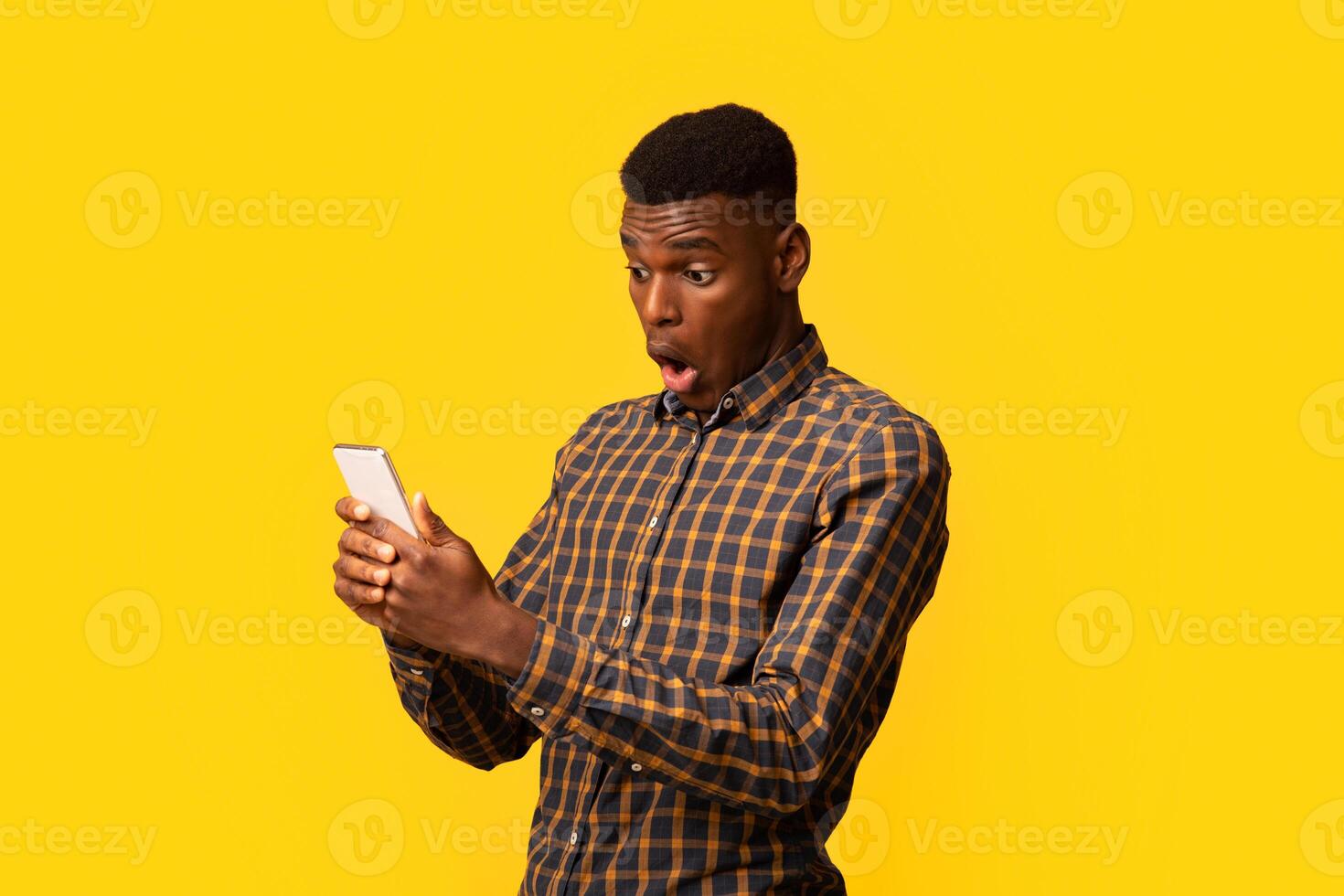 Portraif of shocked funny black man looking at smartphone screen with opened mouth, excited african american guy emotionally reacting to good online offer, standing with isolated on yellow background photo