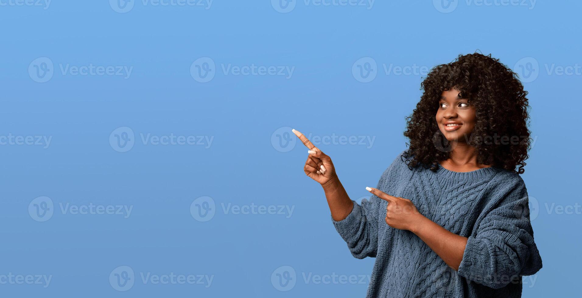 Attractive black young woman in knitted green sweater pointing at copy space and smiling, yellow studio background, panorama. Cute african american curly lady showing empty space for advertising photo