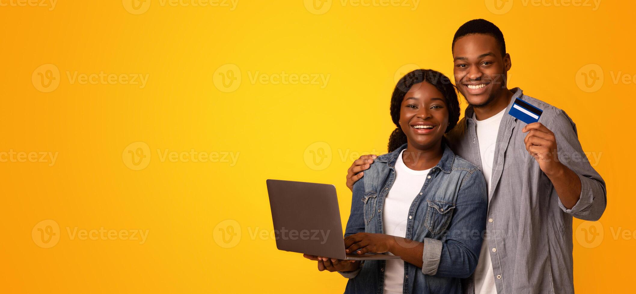 Online Shopping Concept. Portrait of happy african american couple with laptop and credit card posing over yellow background, cheerful black spouses enjoying puchase in internet, free space photo