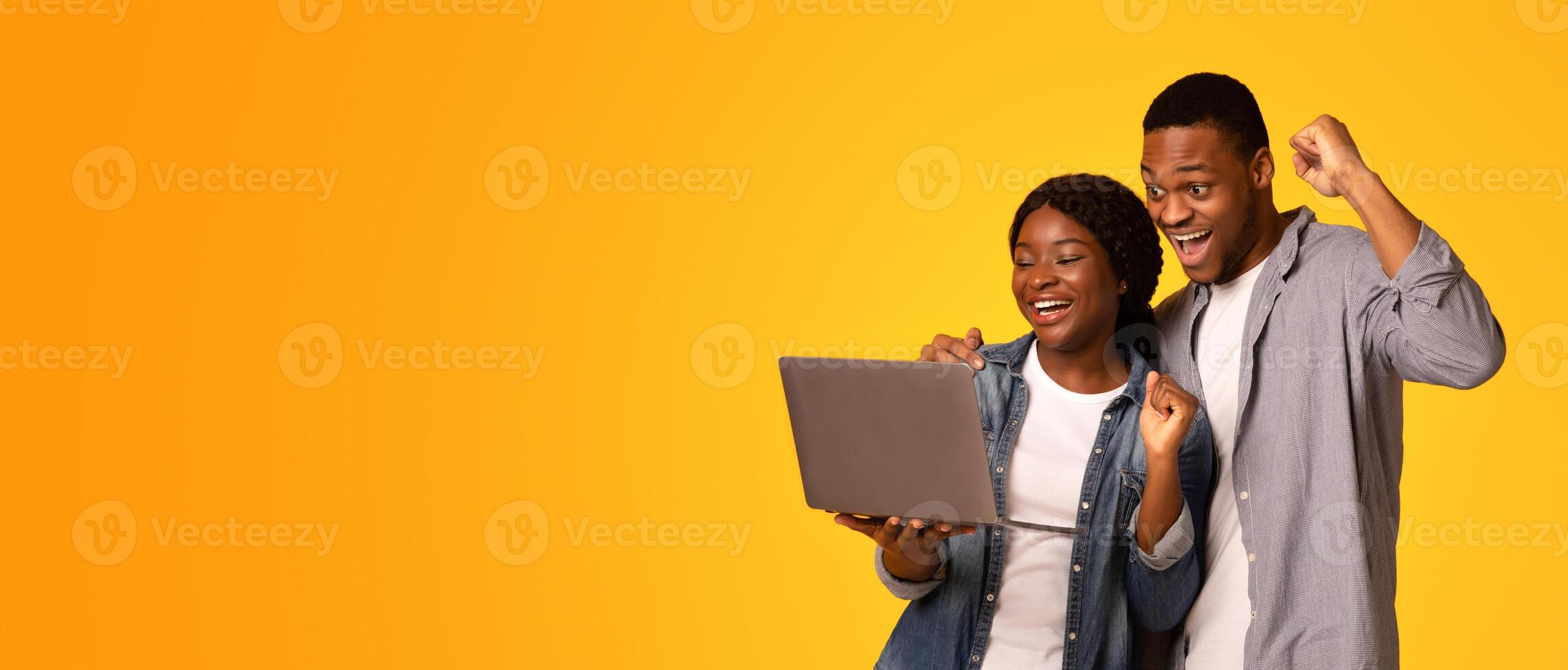 Overjoyed Black Couple Holding Laptop Computer And Raising Fists With Excitement, Emotionally Celebrating Success, Lucky People Standing Isolated Over Yellow Background, Panorama With Copy Space photo