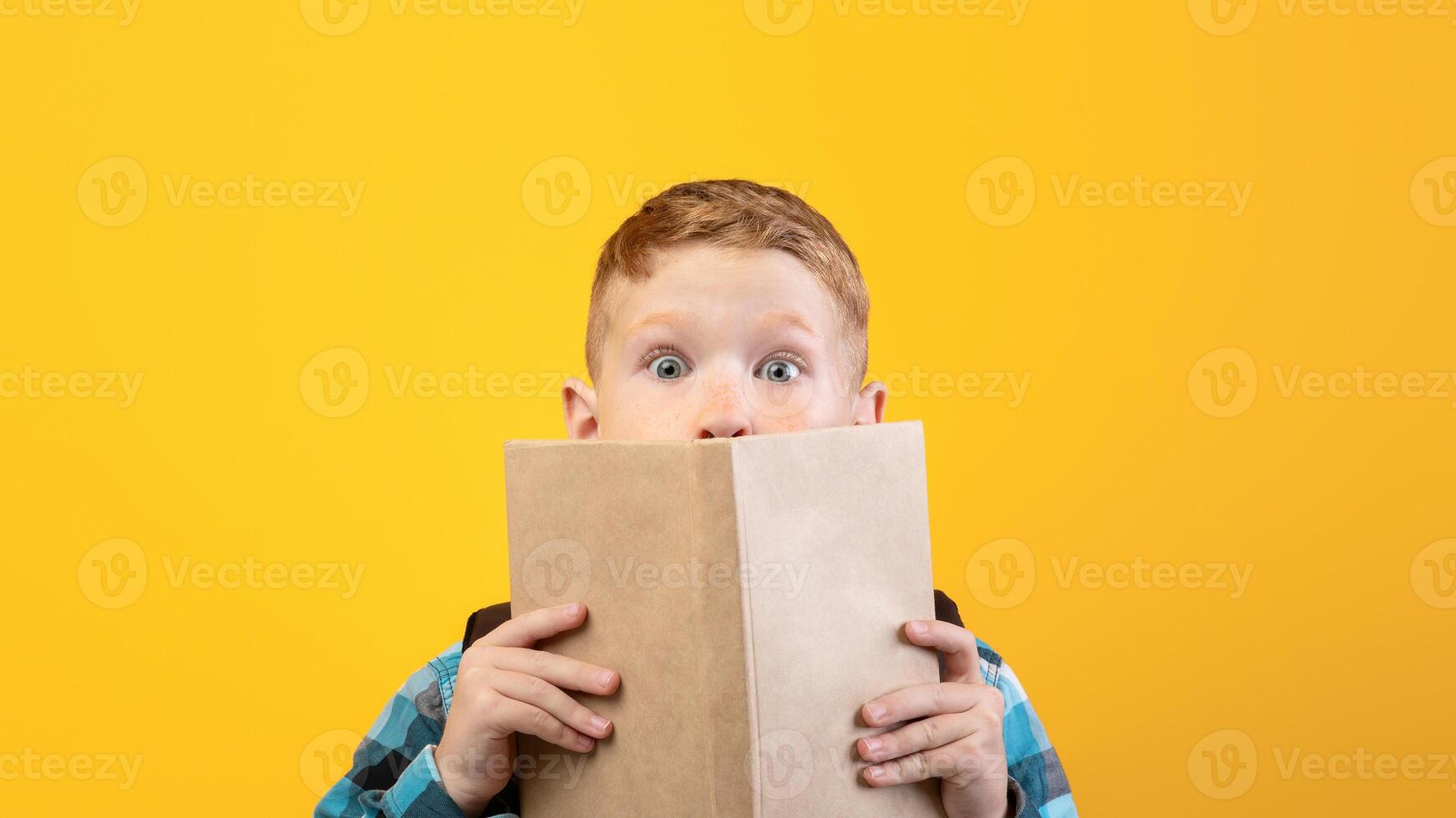 Shocked boy covering his mouth with book, reading exciting stories, yellow studio background, empty space. Emotional smart kid reading book, got impressed by novel. Educational concept photo