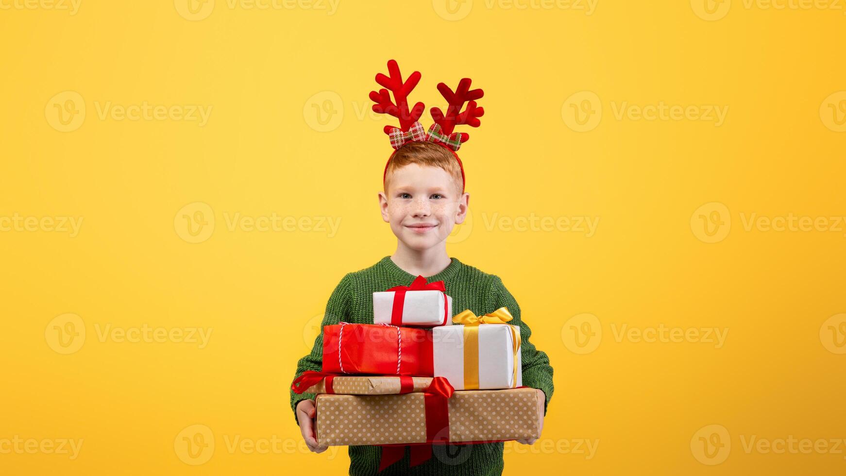 Smiling boy with festive deer accessory holding Christmas gifts over yellow studio background, empty space. Happy kid celebrating New Year, got a lot of presents, cheerfully posing at camera photo