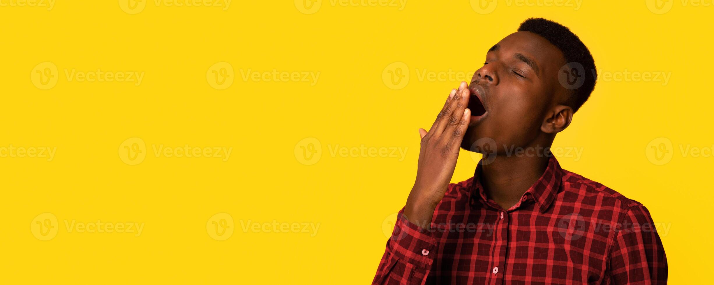 Closeup shot of tired young black man yawning, exhausted african american guy covering opened mouth with hand, feeling bored and sleepy while standing isolated over yellow background photo