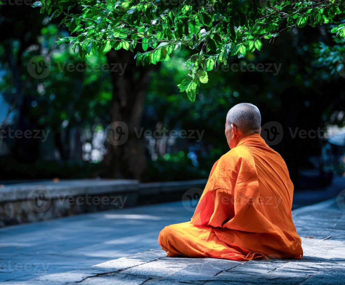 Monk in orange robe meditates in park under tree canopy photo