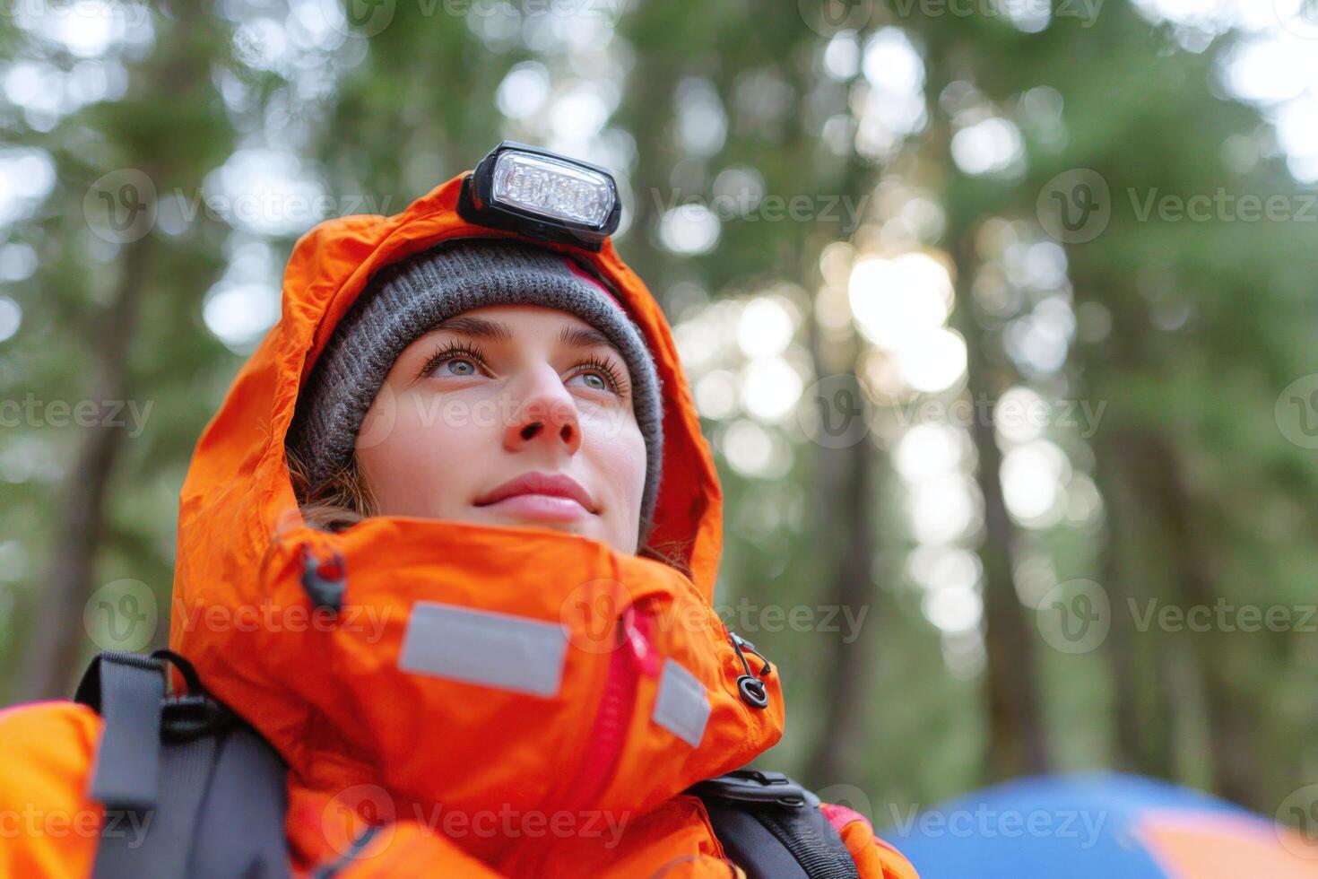 Adventurous woman wearing bright orange outdoor gear and headlamp, gazing upwards in a forest, surrounded by tall trees and a colorful camping tent, embodying the spirit of exploration photo