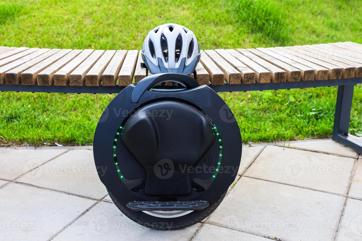 Modern unicycle parked on a wooden bench in a park with green grass and a helmet resting nearby photo