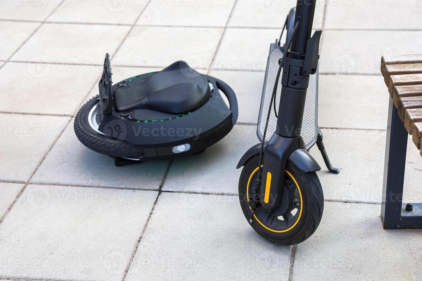 Electric scooter and unicycle parked on a city sidewalk near a wooden bench in bright daylight photo
