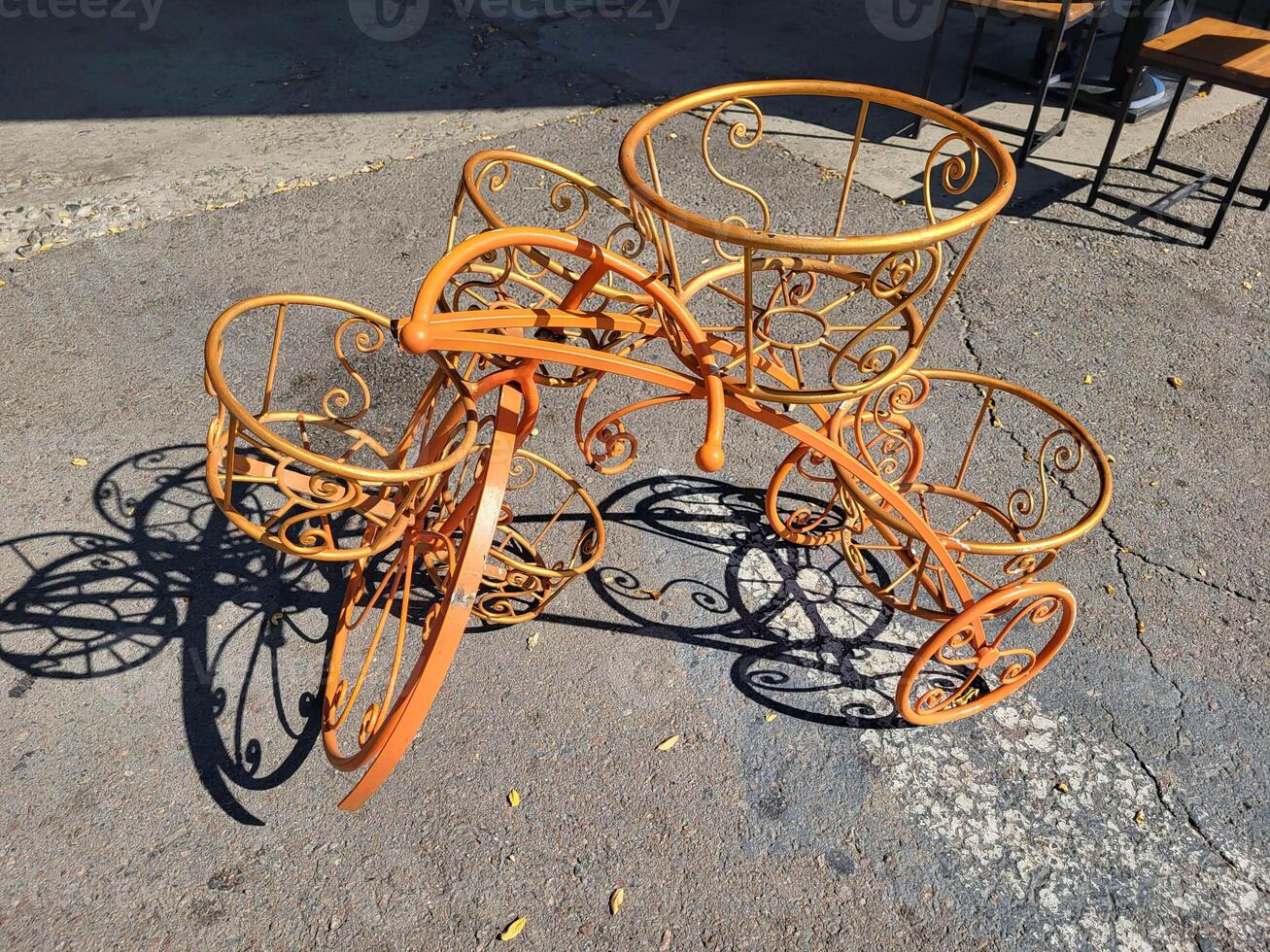 An orange empty decorative wrought-iron flower stand in the shape of a bicycle with baskets for plants, stands on the sidewalk. photo
