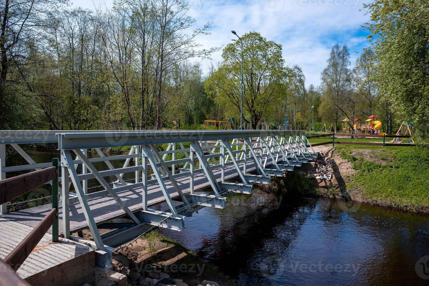 A modern metal pedestrian bridge crosses a calm waterway, connecting grassy areas. A park with slides, swings, and green trees is visible under a sunny sky. photo