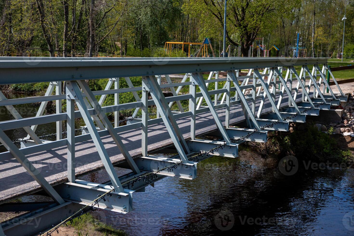 A metal lattice style pedestrian bridge spans a narrow waterway, reflecting in the water. Nearby are trees, a playground with swings, and park features. photo
