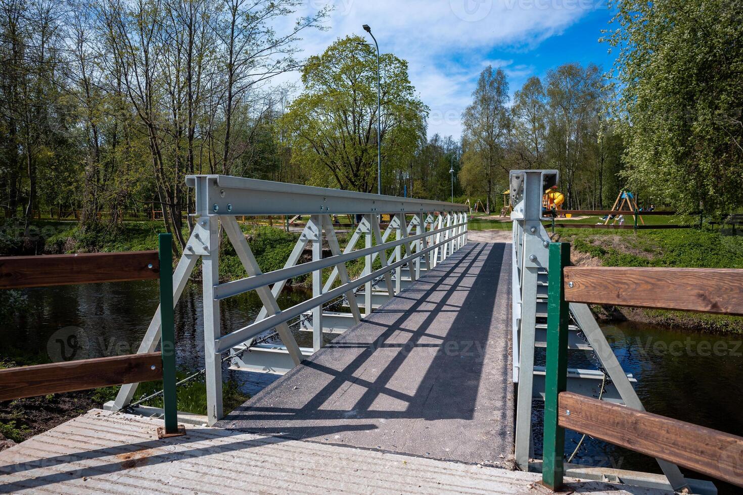 A small pedestrian bridge with crisscrossed metal supports spans a waterway, surrounded by trees and open park space with a playground in the distance. photo