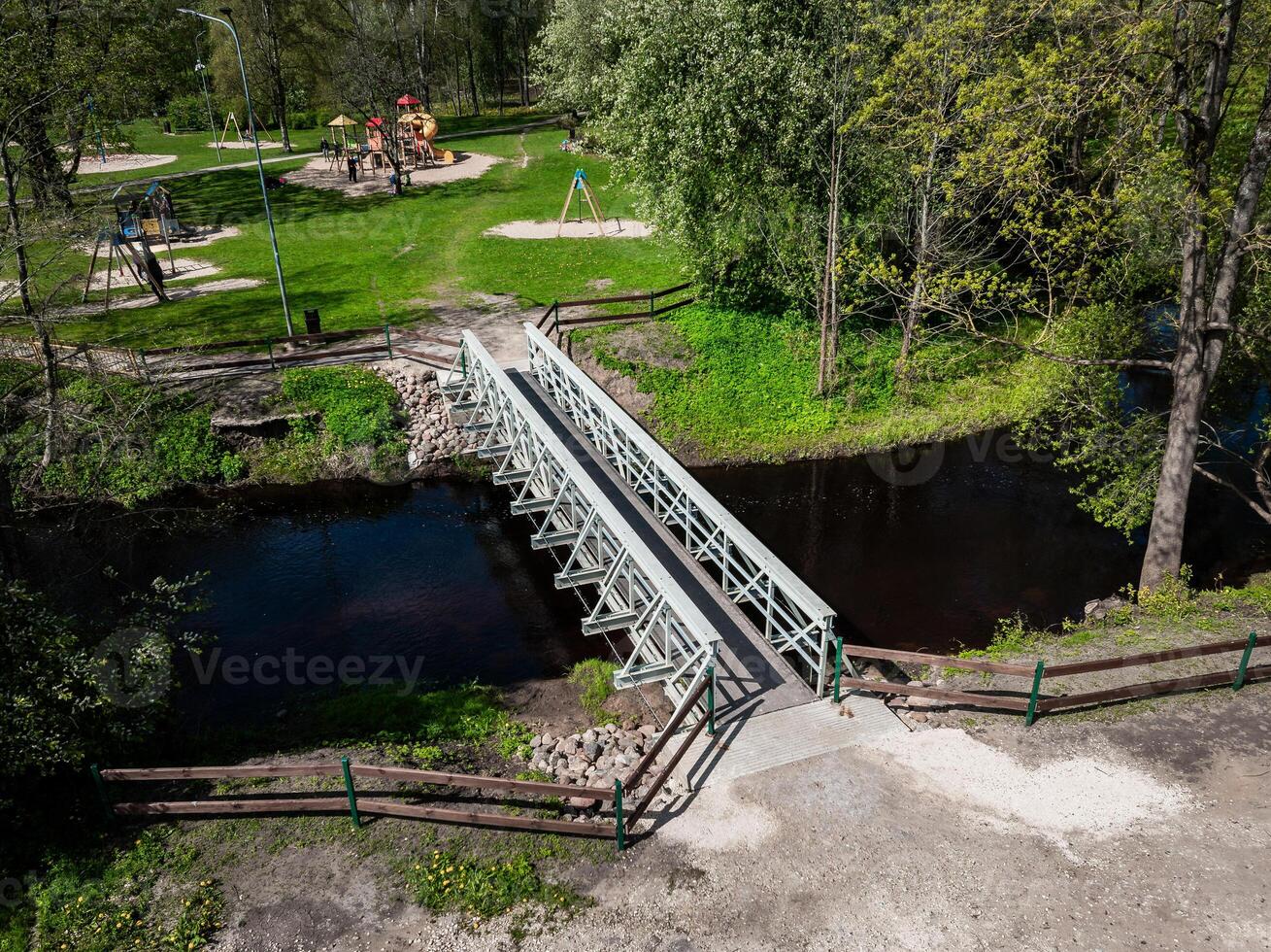 A small metal bridge with wooden railings crosses a narrow stream in a park. The park includes a playground, grassy areas, trees, and visible pathways. photo