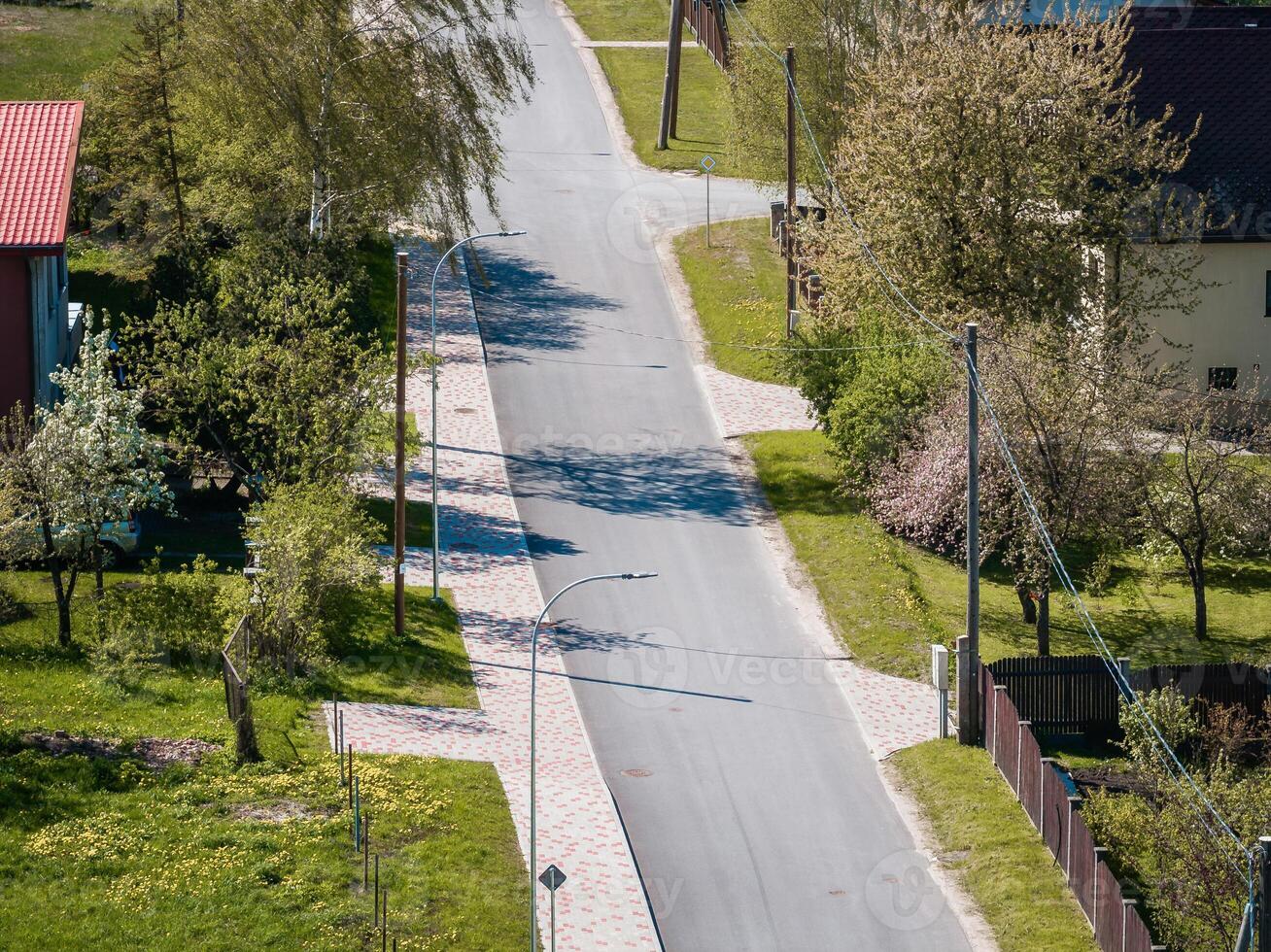 Quiet residential street lined with trees, small houses with red and dark roofs, patterned sidewalks, streetlights, power lines, and blooming gardens. photo