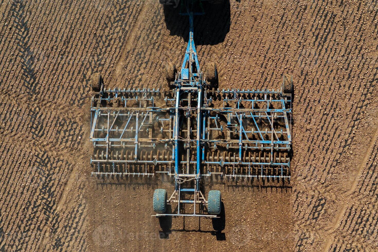 Aerial perspective of blue farming machinery attached to a tractor, tilling a field with evenly furrowed soil and parallel lines visible. photo