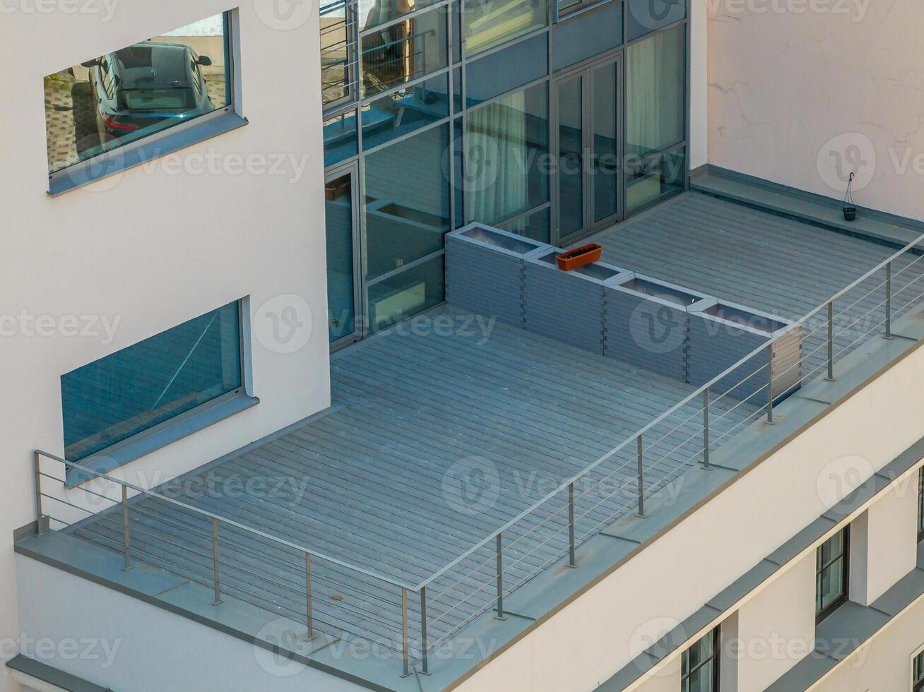 A modern building with a rooftop terrace featuring a wooden deck, metal railing, planter box, large glass windows, and a parked car visible inside. photo