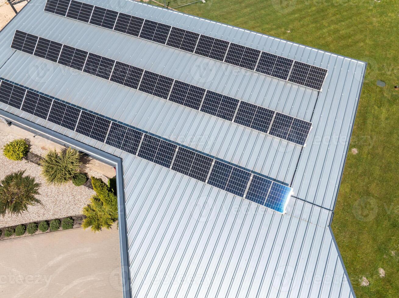 A metal roof with rows of solar panels is surrounded by a gravel area, arranged plants, and a grassy lawn, emphasizing renewable energy design. photo