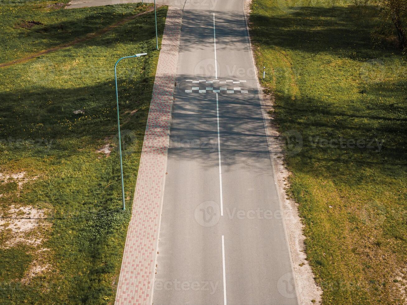 A straight paved road with a marked pedestrian crossing, grass on both sides, a streetlamp on the left, and a brick paved sidewalk parallel to the road. photo
