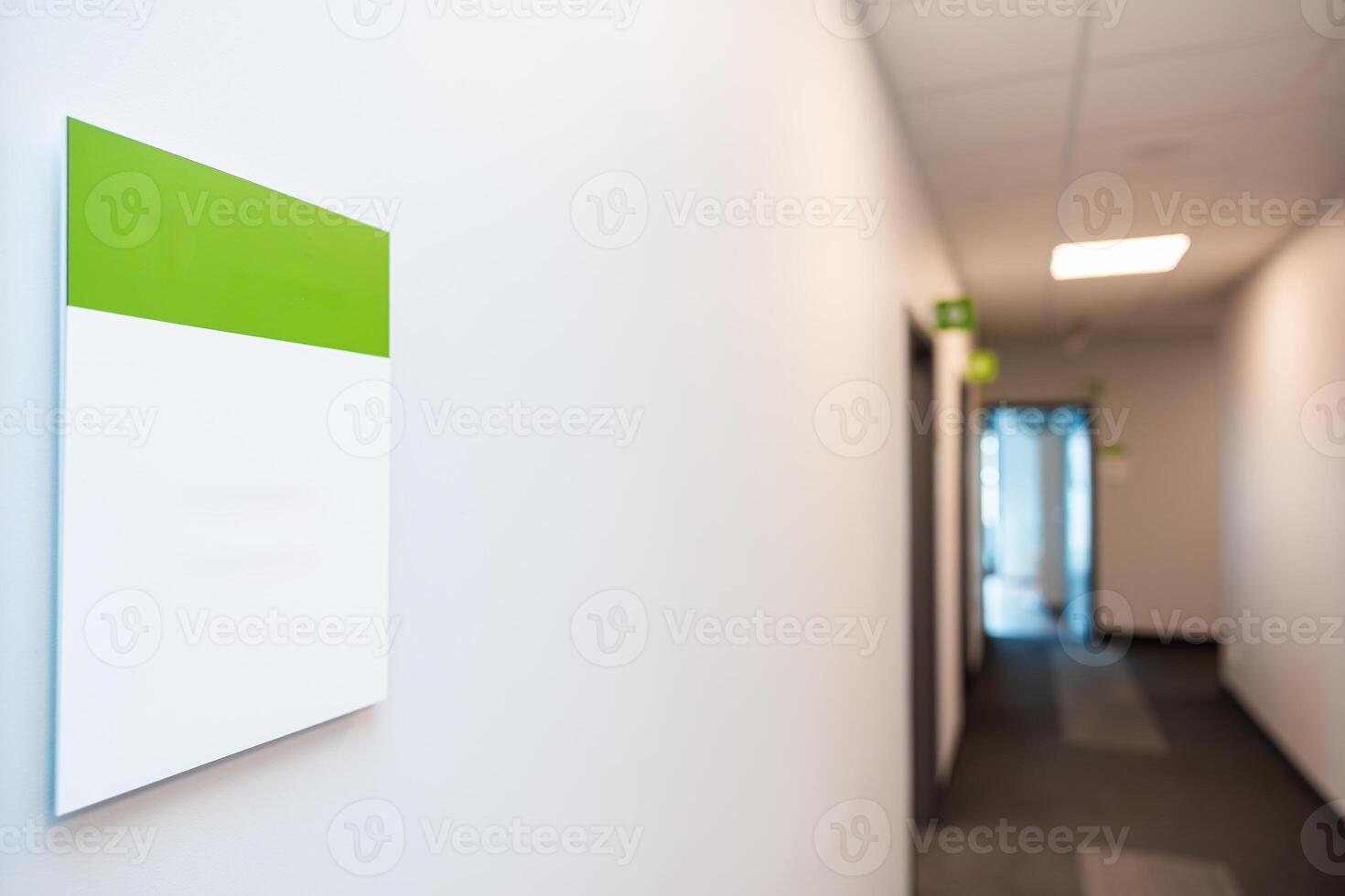 Well lit hallway with white walls, green and white sign, carpeted floor, multiple doorways, and a glass door at the end allowing natural light. photo