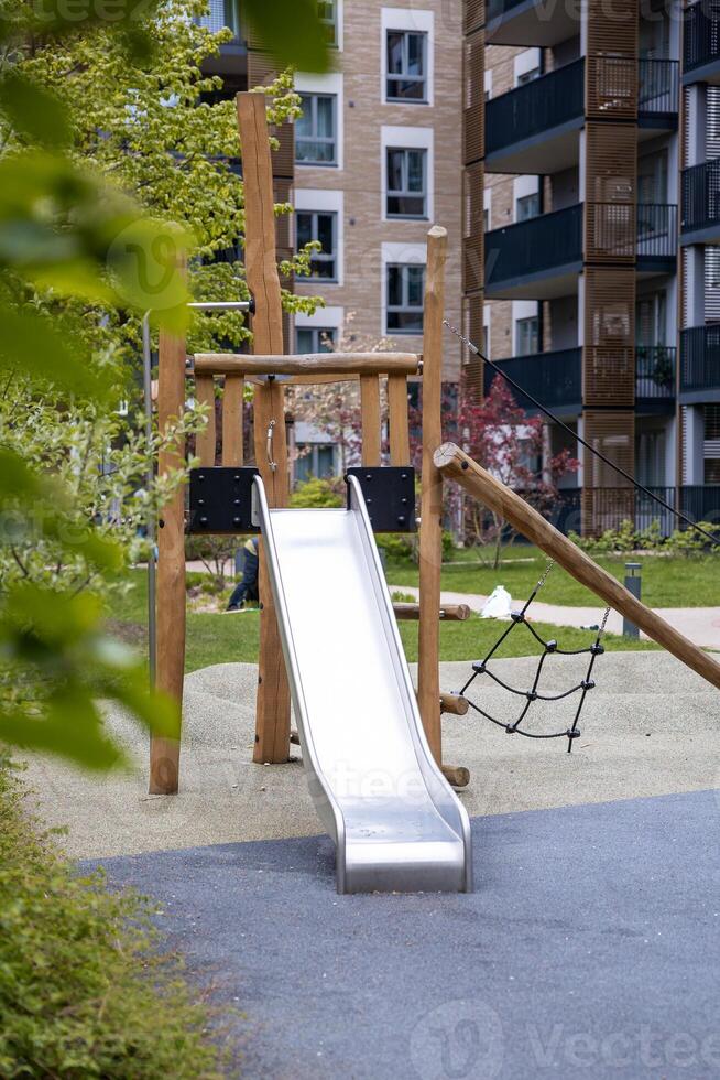 Modern playground featuring a metal slide, wooden frame, and climbing net, surrounded by pathways, greenery, and multi story apartment buildings. photo