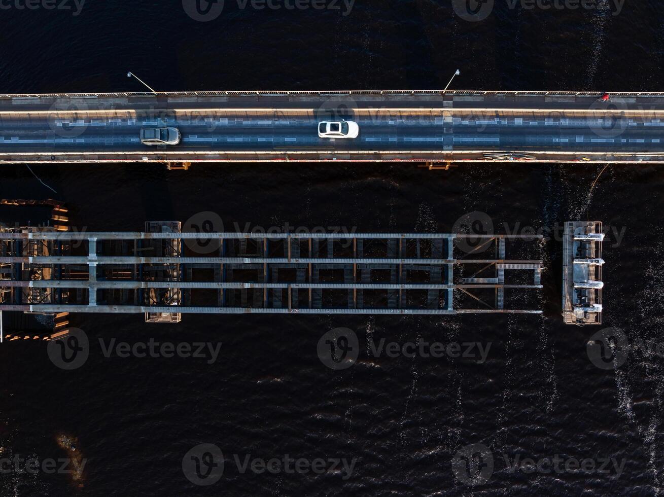 An aerial view shows a drawbridge with one section raised, vehicles on the roadway, a person in red walking, and dark water beneath the structure. photo