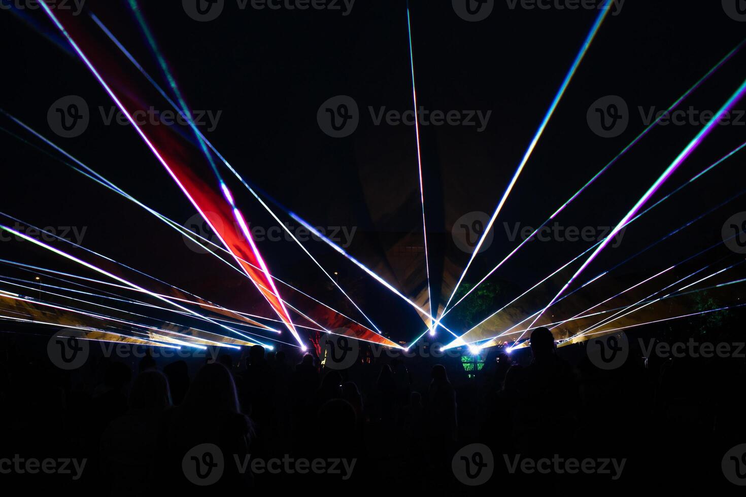Vibrant laser beams form dynamic patterns in the dark night, with silhouetted people in the foreground and faint structures or trees in the distance. photo