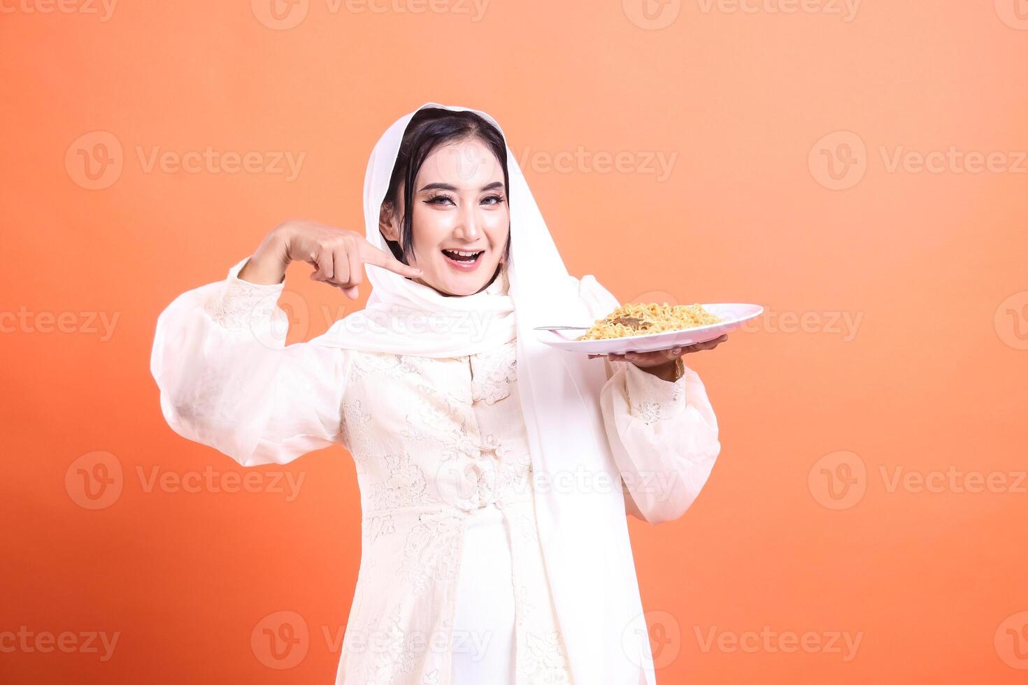 Smiling Muslim woman in a hijab holding and offering a plate of noodles, with a happy expression and pointing with one finger. isolated on a orange background photo