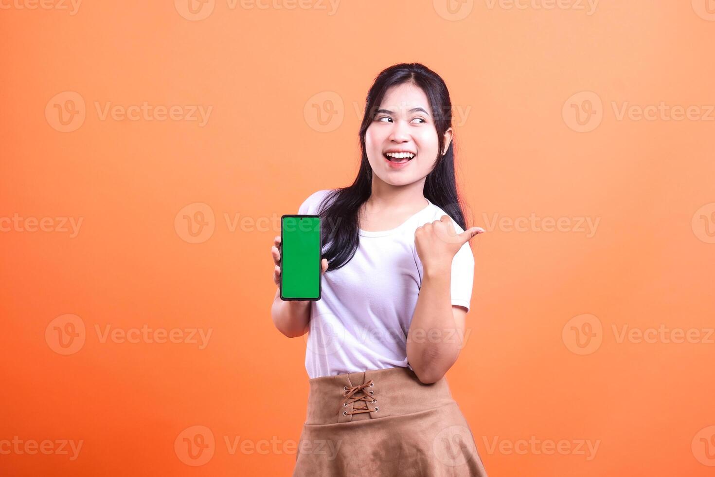 Cheerful woman in white t-shirt presenting green screen phone pointing to the side on orange background photo