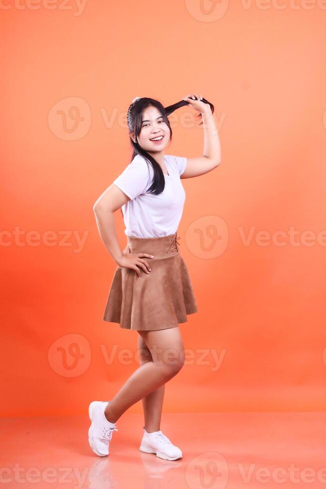 A cheerful young woman in a white t-shirt and brown skirt playfully holds her hair isolated on orange background photo