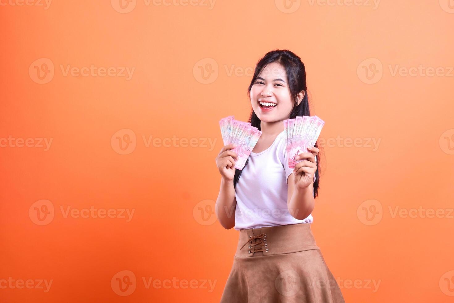 Smiling woman holding a lot of money in both of her hands standing against orange background photo