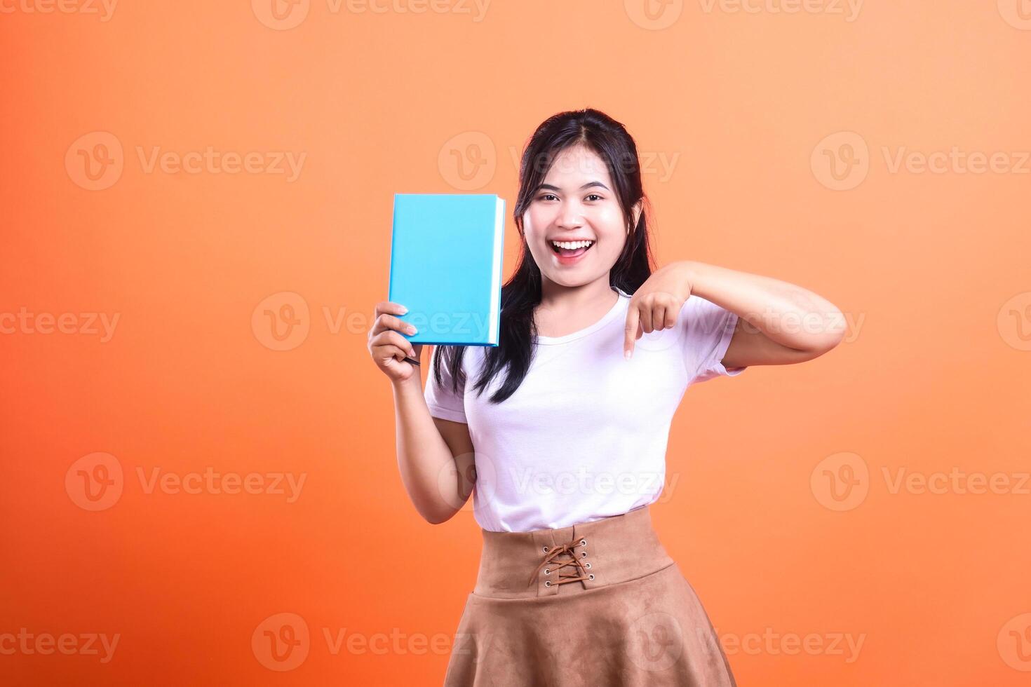 A woman holding a blue book, points down with her right hand, wearing a white shirt and brown skirt isolated on orange background photo