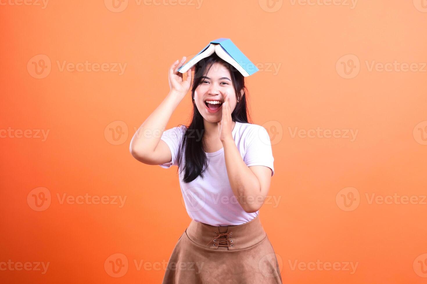 A smiling woman with a book on her head and hand on her mouth stands against an orange background photo