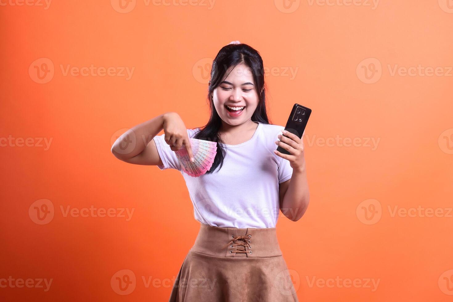 Excited Woman Holding Indonesian Rupiah and smart phone device Against Orange Background photo
