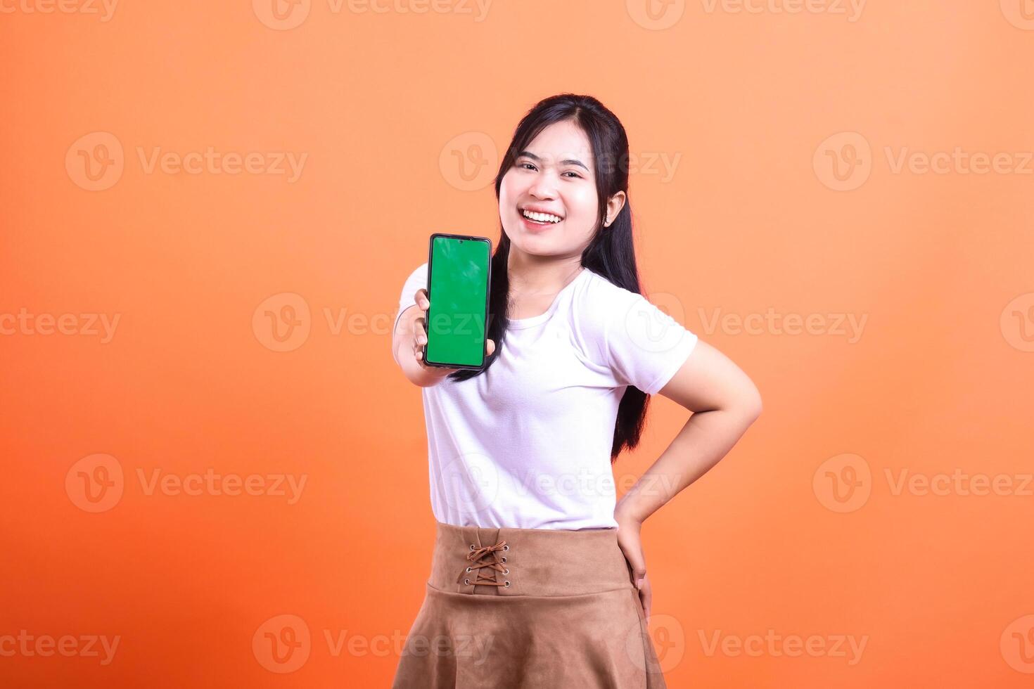 Smiling woman holding out mobile phone with green screen in front of orange backdrop photo