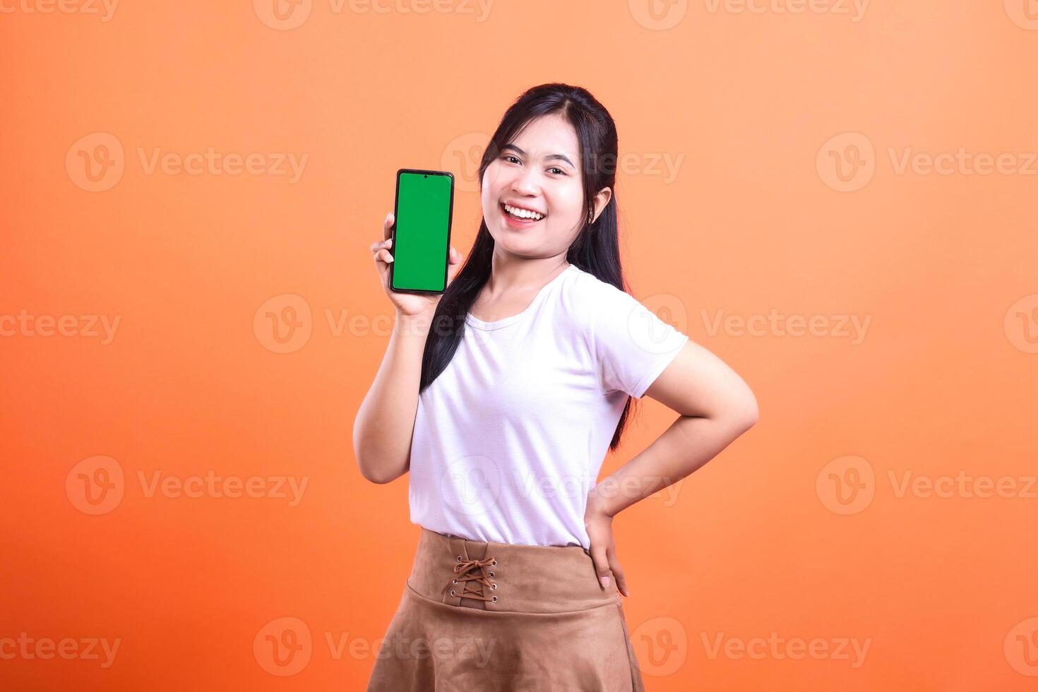A cheerful woman presents a smartphone with a green screen against orange background photo