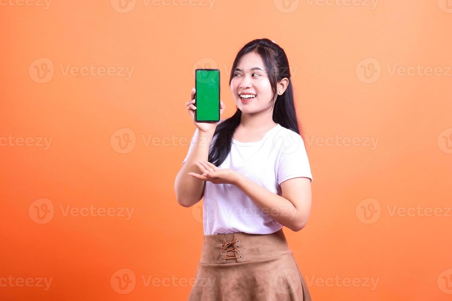 Smiling Woman Presenting Smartphone with Green Screen on Orange Background photo