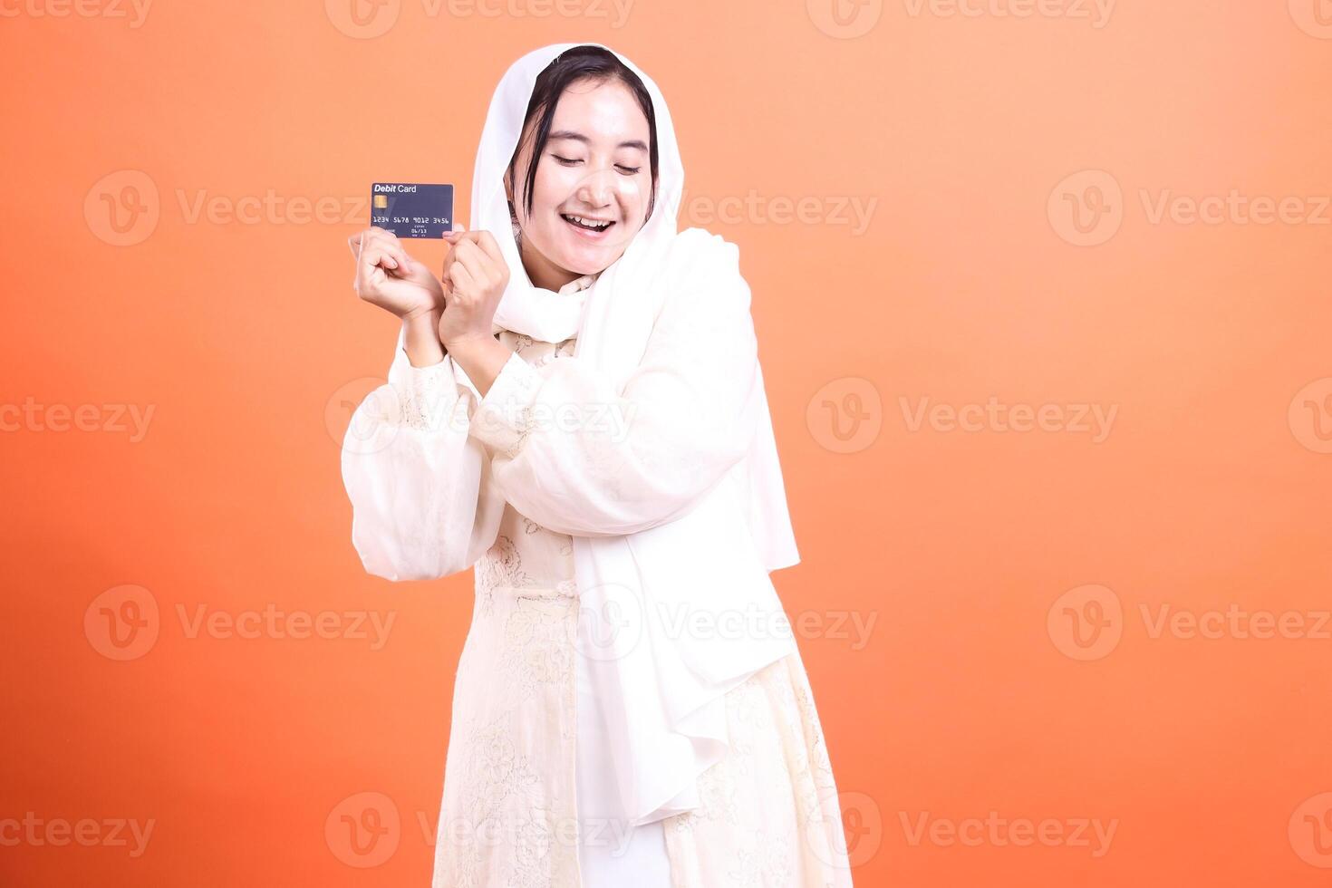 Cheerful Muslim woman with debit card She is incredibly happy to have a debit card in her hand, isolated on a orange background. photo