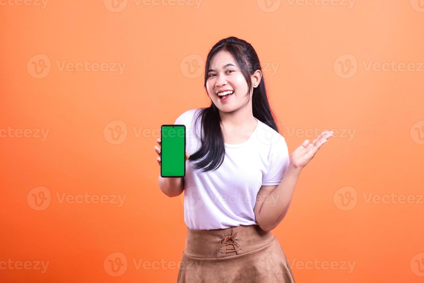A woman holds a smart phone gadget with a green screen isolated on an orange background. She smiles and gestures as if she is presenting or demonstrating something on the phone. photo
