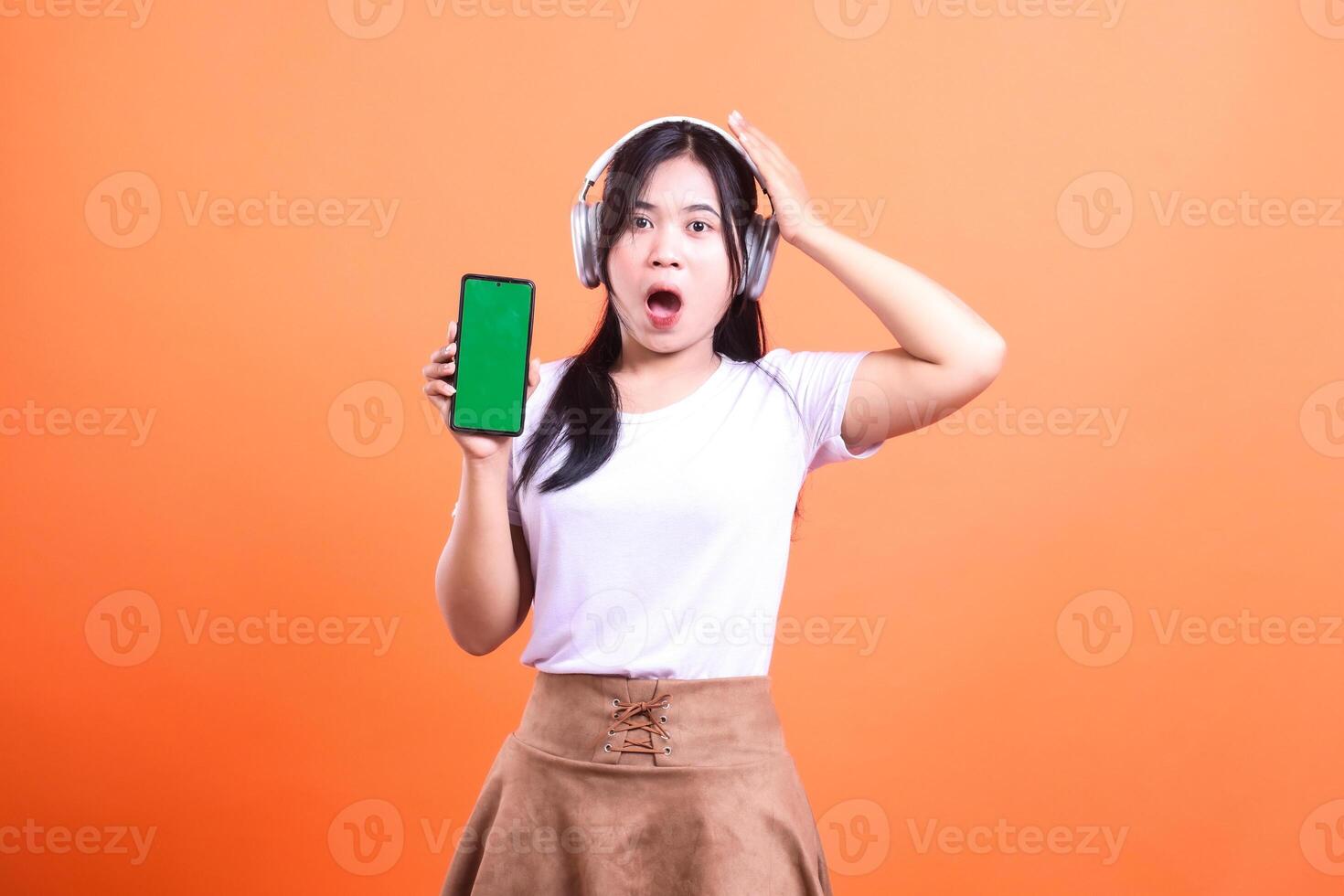 Woman holding a green screen smart phone gadget with headphone with shocked expression in an orange background photo