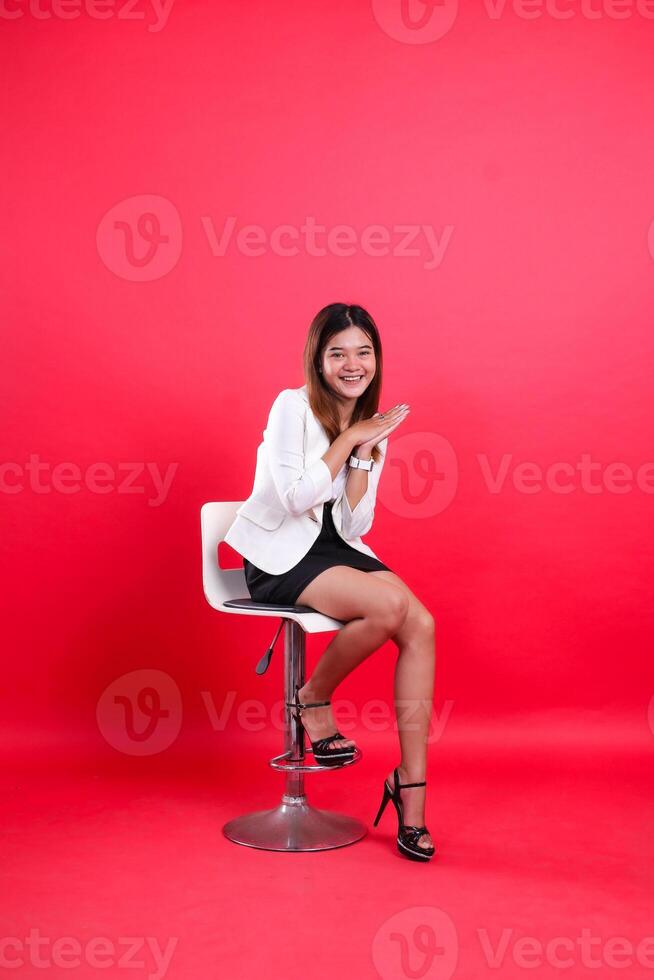 Full body young Asian woman in white coat and shorts, cheerful to camera, hands together, sitting on hydraulic chair isolated on red background photo