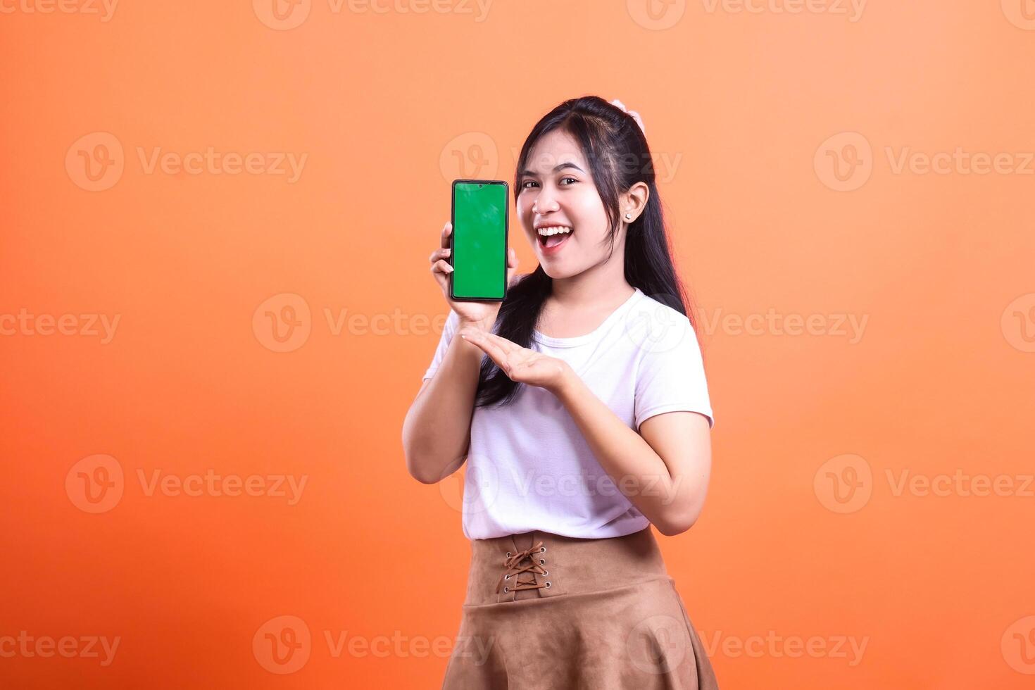 Young Woman Presenting a Smartphone with Green Screen on Orange Background photo