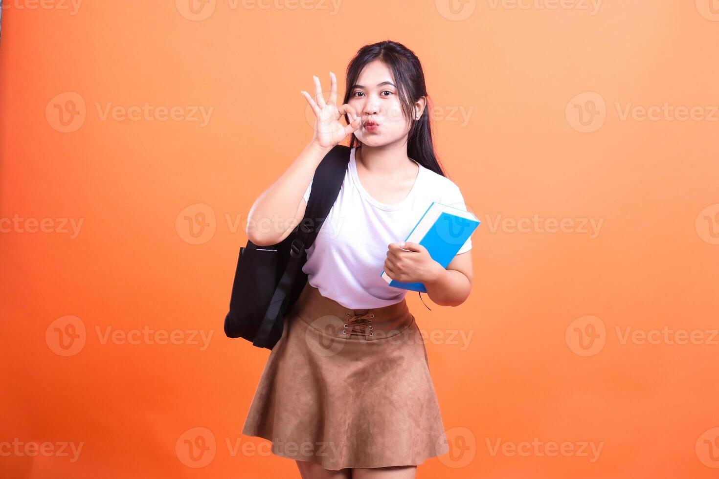 Young female student poses with a book and backpack with OK gesture in front of orange backdrop photo