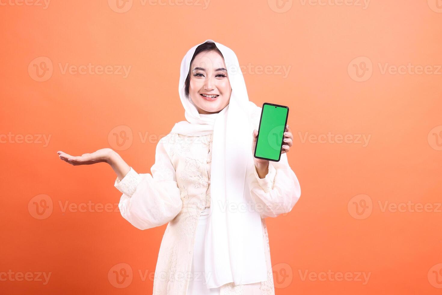 Smiling muslim woman in hijab holding up smartphone with green screen and presenting with hand gesture for copyspace. isolated on a orange background. photo